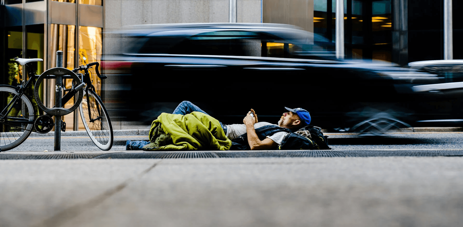 A man lies on the sidewalk in an urban setting, wrapped in a green sleeping bag and holding a phone, while blurred cars rush past him on the street. A bicycle is locked to a post nearby, and tall office buildings reflect the bustling city life around him.