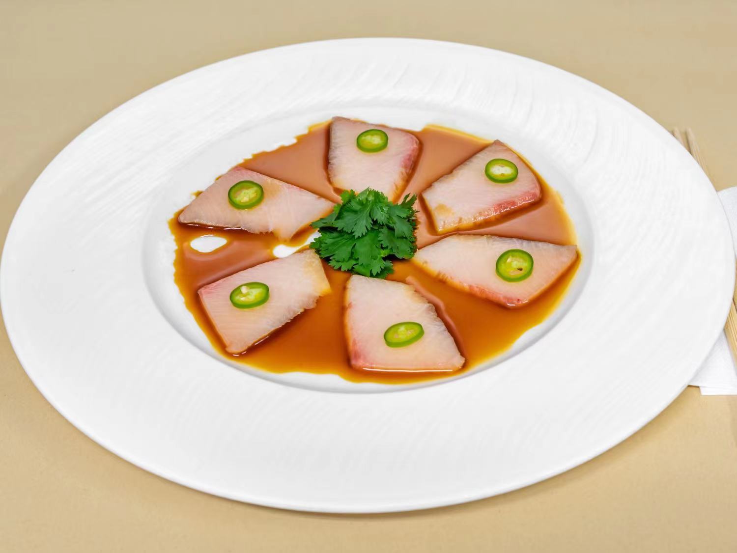 Thinly sliced yellowtail sashimi arranged in a circular pattern, topped with a slice of jalapeño on each piece and drizzled with ponzu sauce. Garnished with a fresh herb in the center, this dish balances rich, fresh fish with a zesty kick.