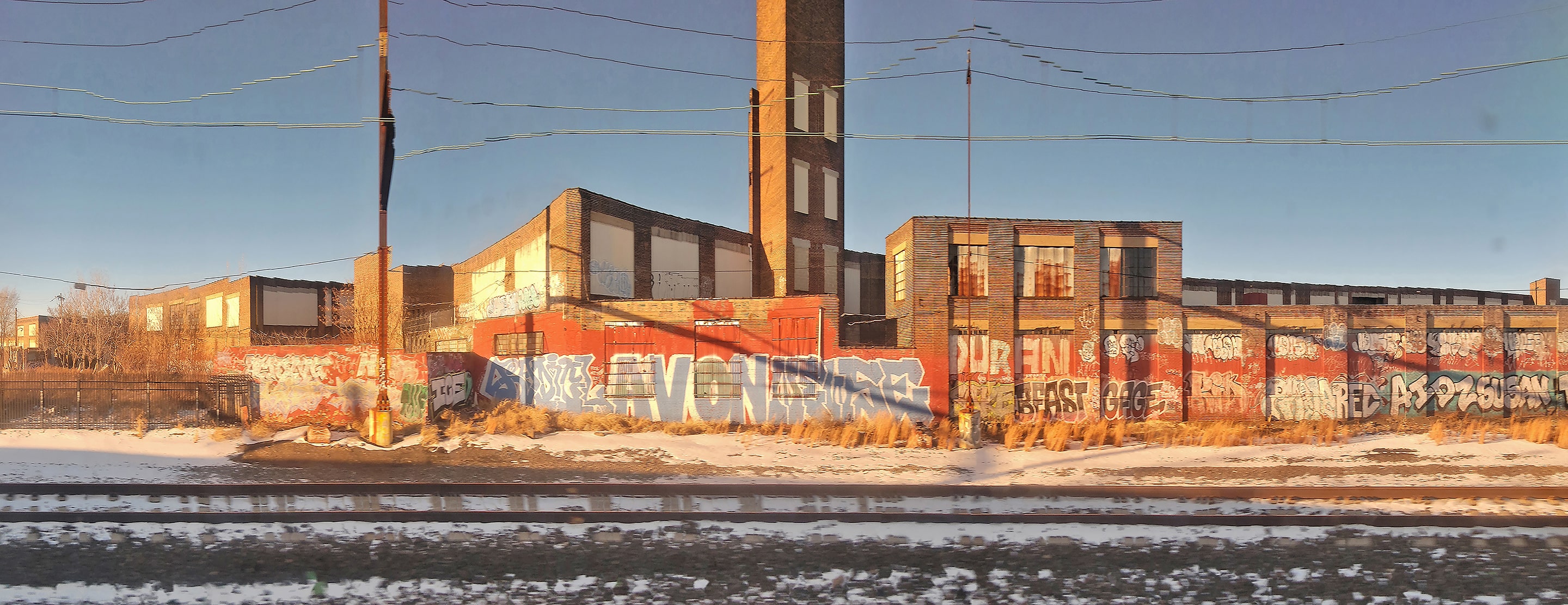 Panoramic photograph of an abandoned industrial building covered in graffiti, with train tracks and patches of snow in the foreground. The warm sunlight and subtle distortions across the frame evoke both decay and quiet resilience.