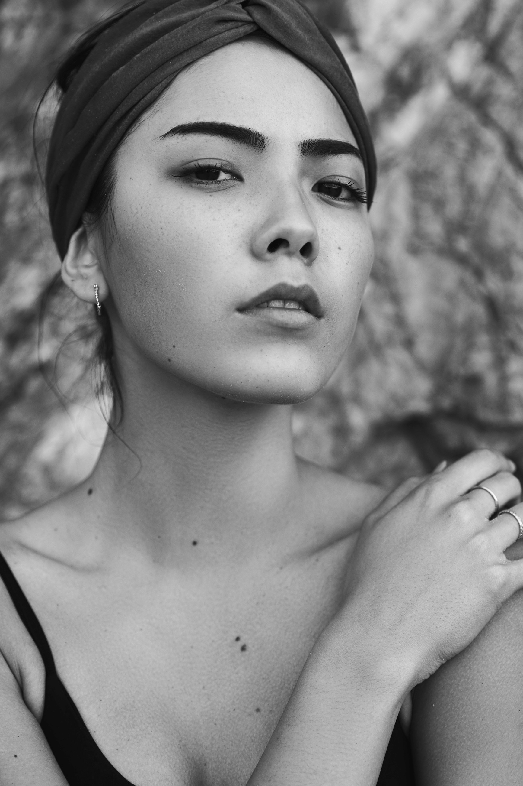 A black and white portrait of an Asian model wearing a headscarf tied at the front and looking at camera with textured rock in the background.
