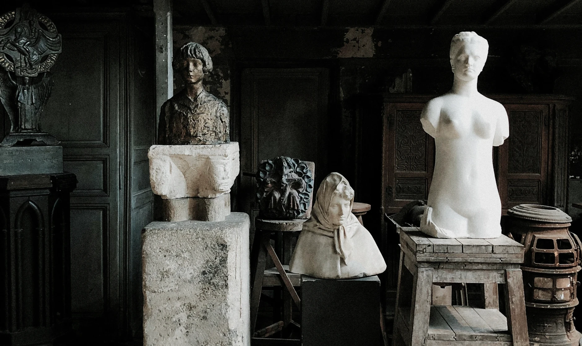 Collection of weathered sculptures and busts in a dimly lit atelier, blending marble and bronze textures that evoke timeless artistry and quiet reverence.