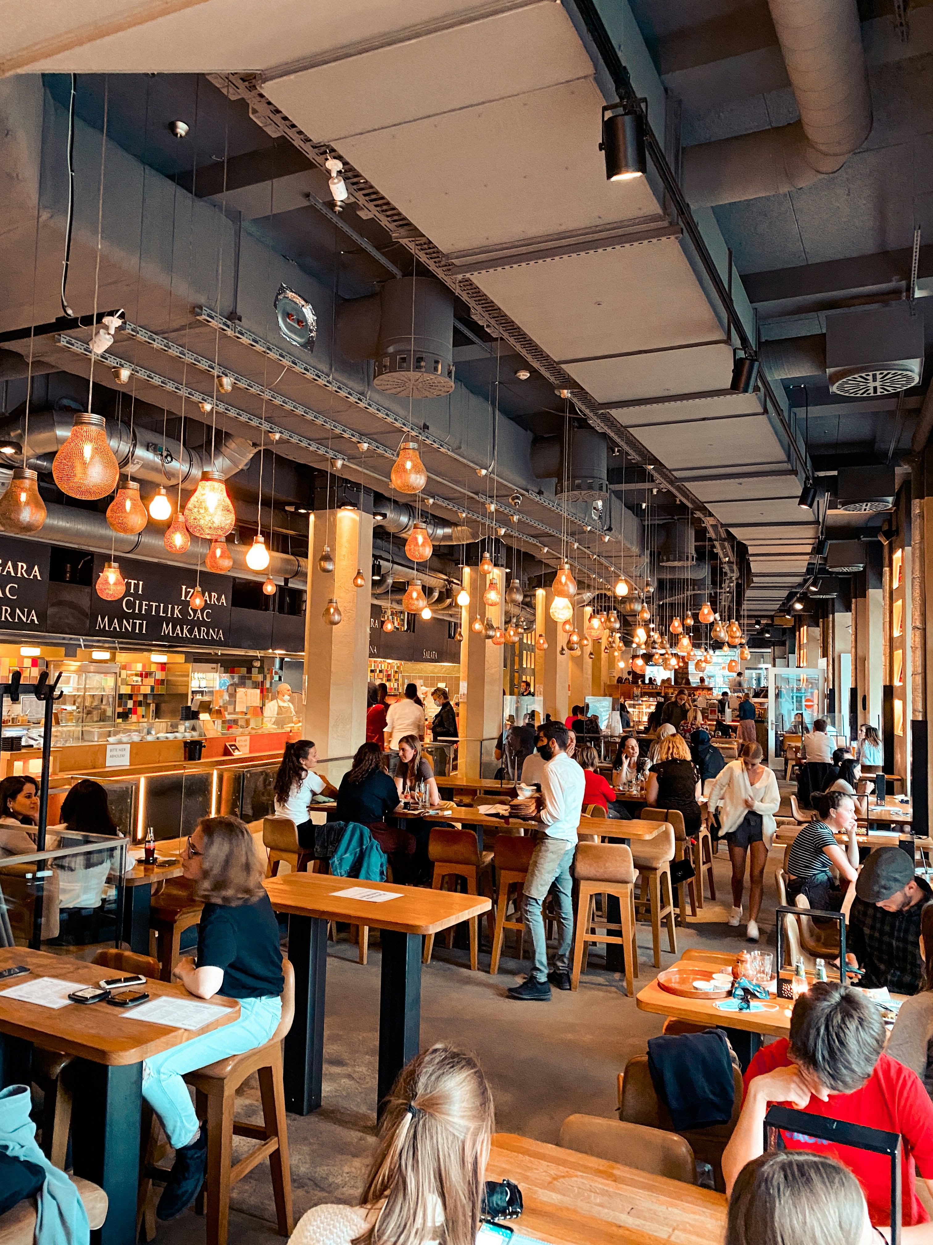 Una escena de calle concurrida con personas reunidas afuera de una tienda o café. El letrero sobre la tienda dice "EIS", lo que sugiere que podría ser una heladería o una tienda de postres. El entorno parece una zona urbana abarrotada.