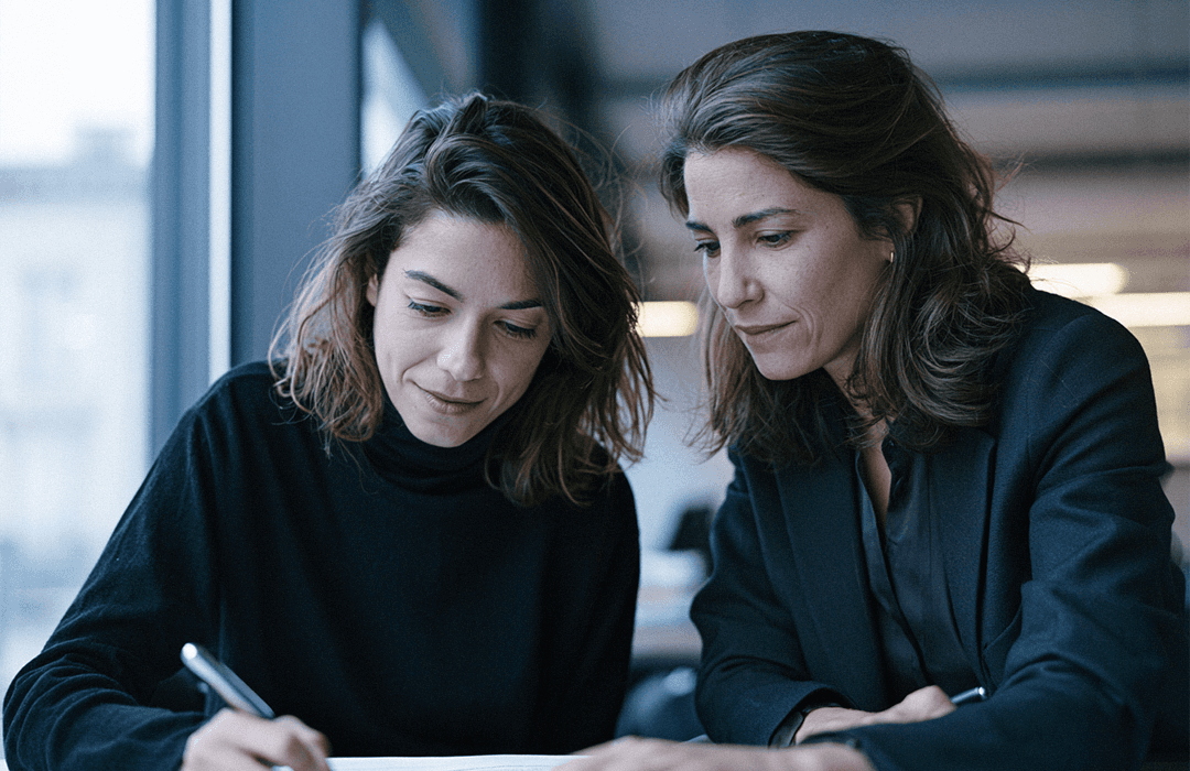 Duas mulheres de terno analisando e assinando documentos juntos em uma mesa de escritório.