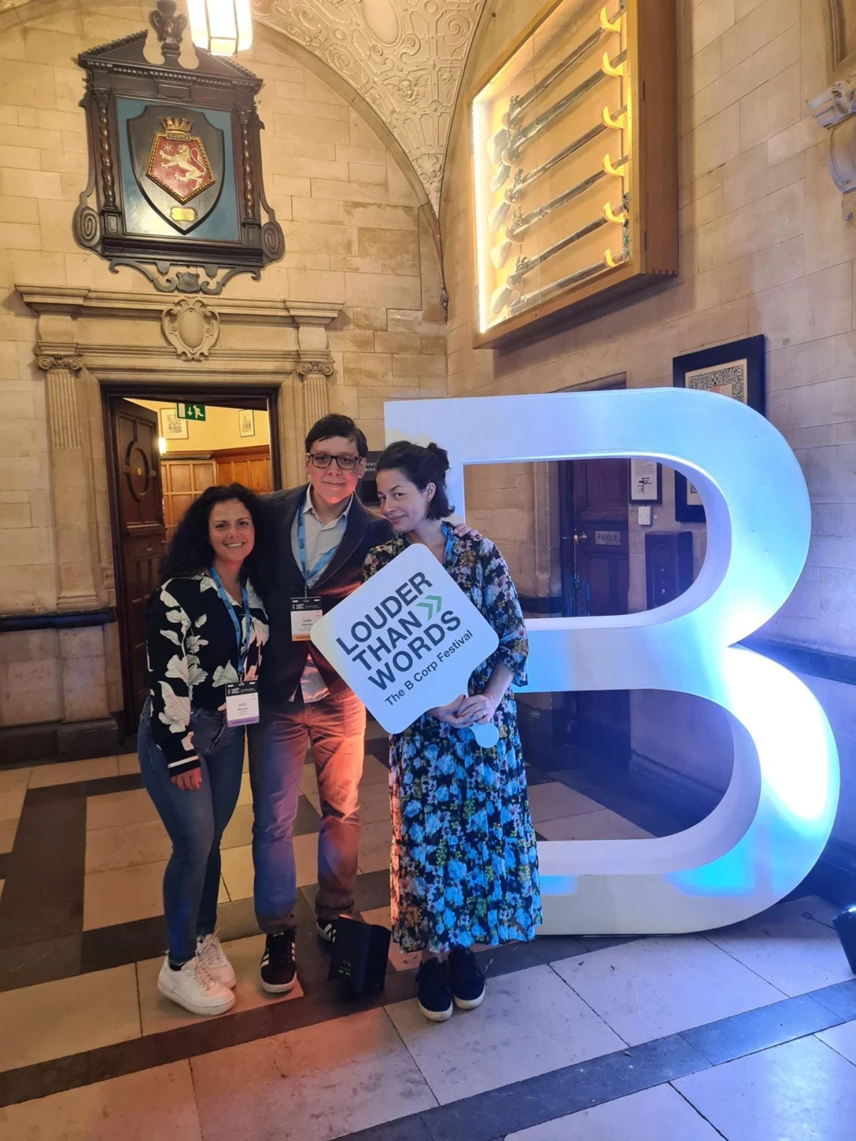 Three people stand together in an ornate hall featuring stone walls, carved details, and a display of historic swords. They pose beside a large illuminated letter “B,” with one person holding a sign that reads “LOUDER THAN WORDS – The CIPD Awards.”