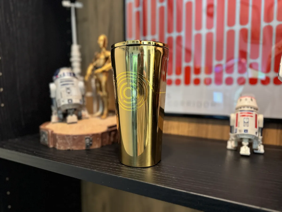 A Corkcicle tumbler with a golden C3PO design on a dark booshelf with a modle of C3PO and R2D2 in the background and a figure of R5-D4 on the other side