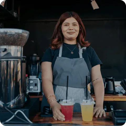 A barista with red hair wearing a gray apron stands behind a counter in a coffee shop, holding two colorful drinks in plastic cups with black straws, surrounded by coffee equipment.