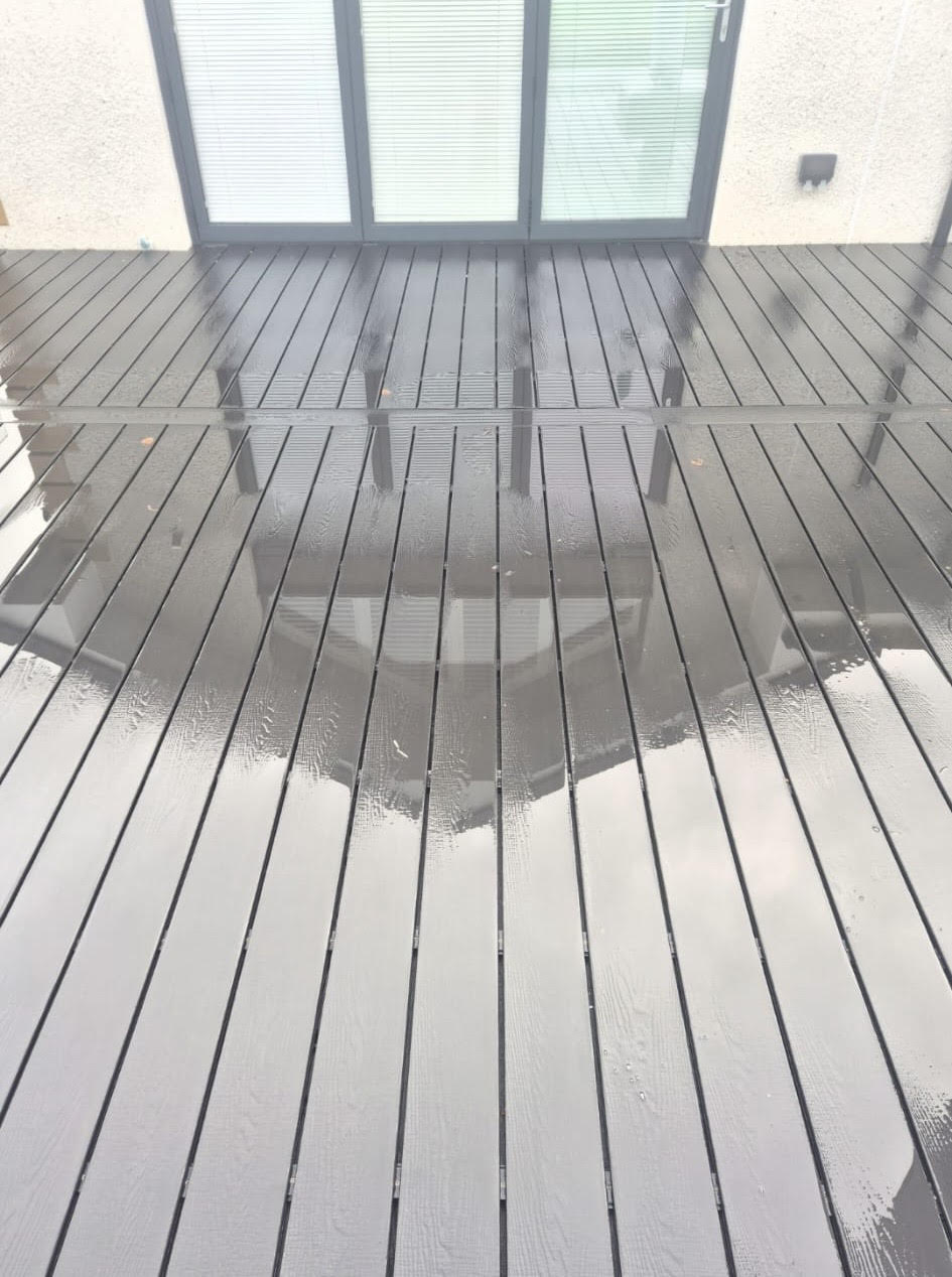 Wet wooden deck reflecting the image of glass patio doors and cloudy sky. The surface appears glossy and polished.