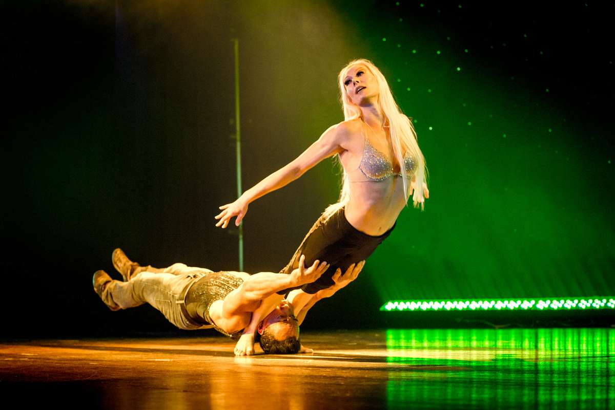 A blonde woman in a sequined top and shorts performs on stage with a man lying on the floor beneath her under green lights.
