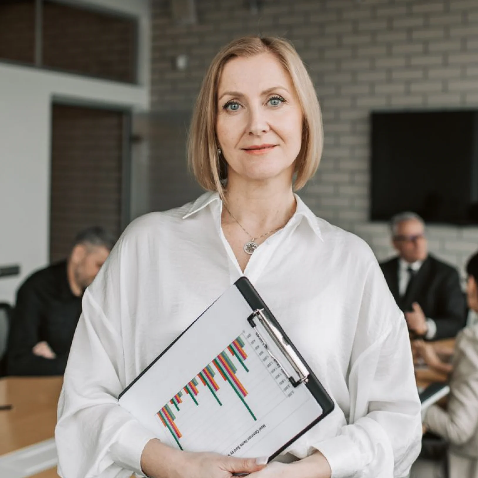 A middle-aged woman with blonde bob-cut hair standing and looking directly at the camera with a gentle smile in an office setting. She is wearing an elegant white long-sleeved button-down shirt and a delicate necklace with a circular pendant. She holds a black clipboard against her chest, displaying a sheet of paper with colorful bar charts. In the blurred background, colleagues in business attire are seated around a conference table for a meeting.
