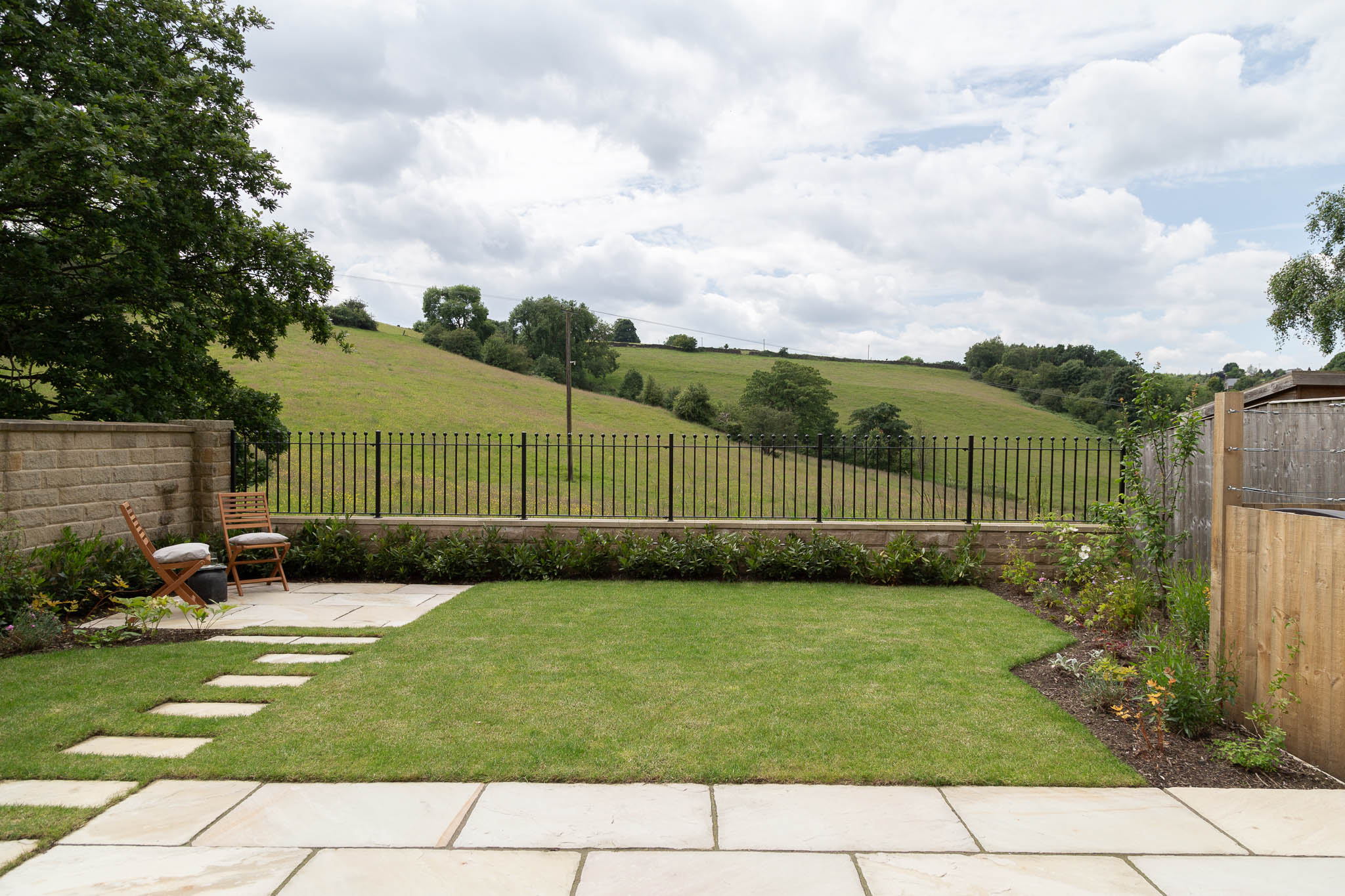 A modern outdoor patio features a sleek, black metal pergola with a built-in grill, surrounded by lush green planting with glimpse views to a cedar clad garden room, offering a stylish and serene garden oasis.