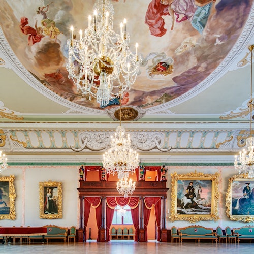 装饰华丽的舞厅配有吊灯、大型画作、红色帷幕的舞台，以及描绘天空中人物的天花板壁画。