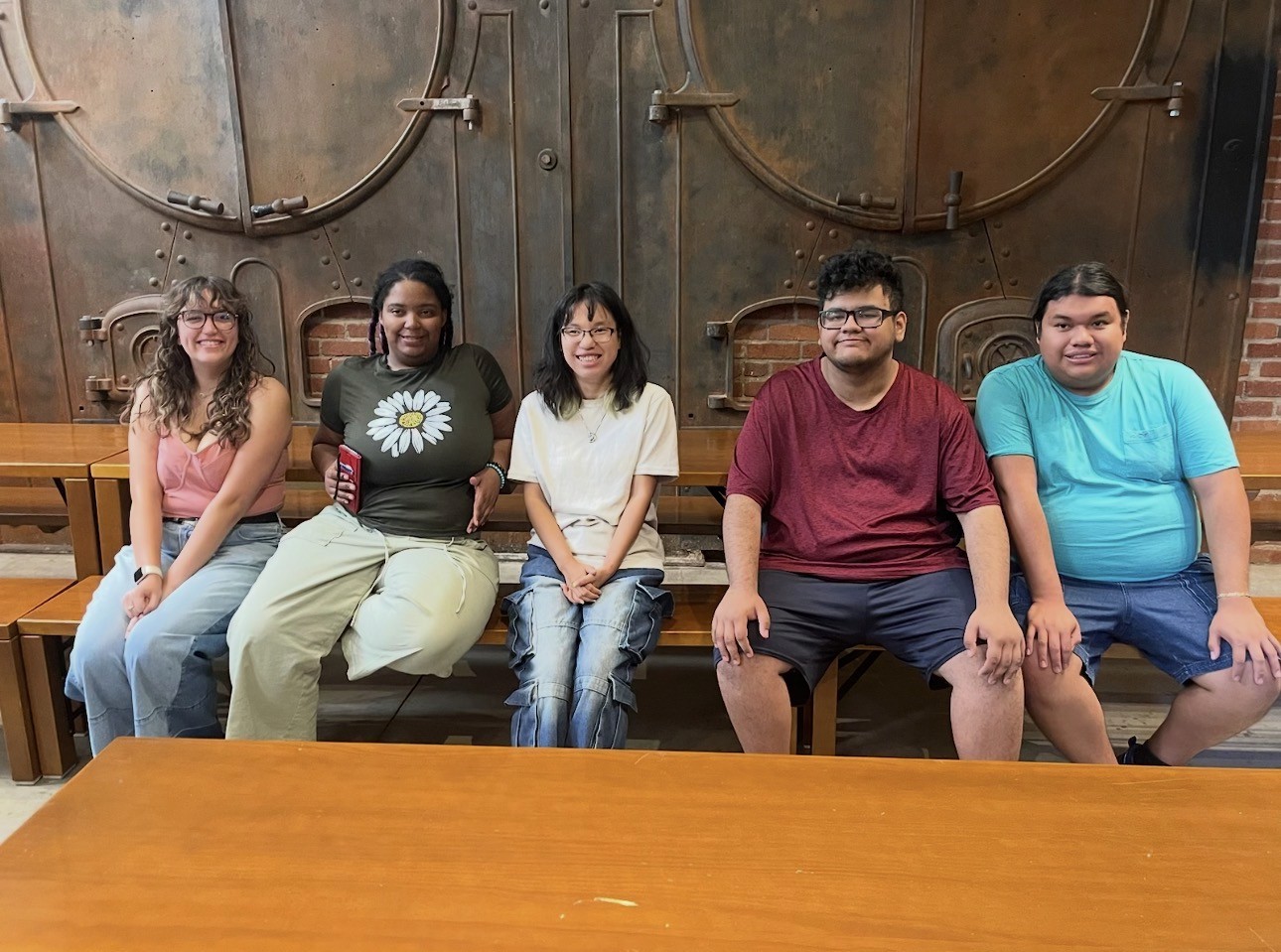 Five Creative Careers interns sit together on a wooden bench in front of large industrial metal doors at the Denver Art Museum.