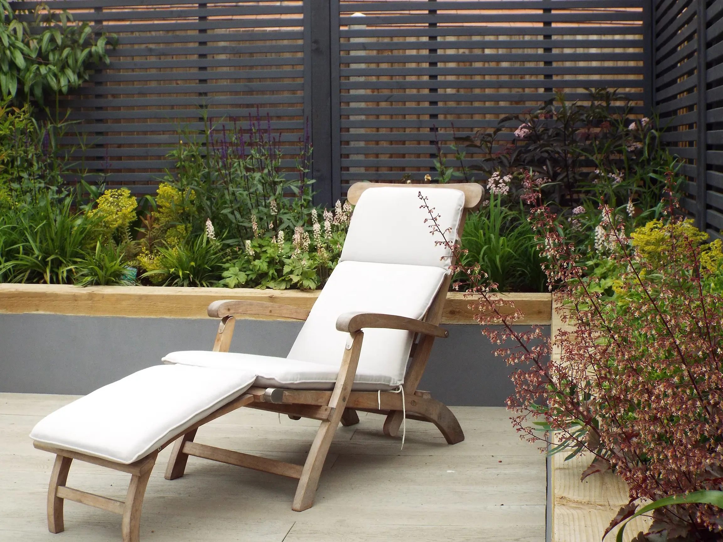 A wooden lounge chair rests in a garden, surrounded by greenery and a stylish outdoor setting.