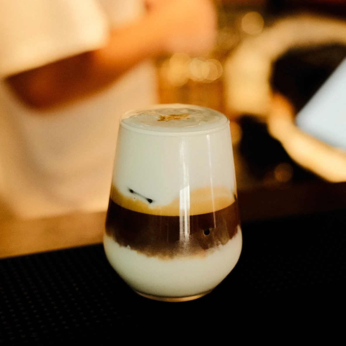 צילום תקריב אסתטי של כוס קפה קרה עם שכבות ברורות של חלב וקפה, מוצבת על דלפק בר כהה. צילום מזון מקצועי עם עומק שדה רדוד מאת Shiriz הצלמת שירי עציץ.