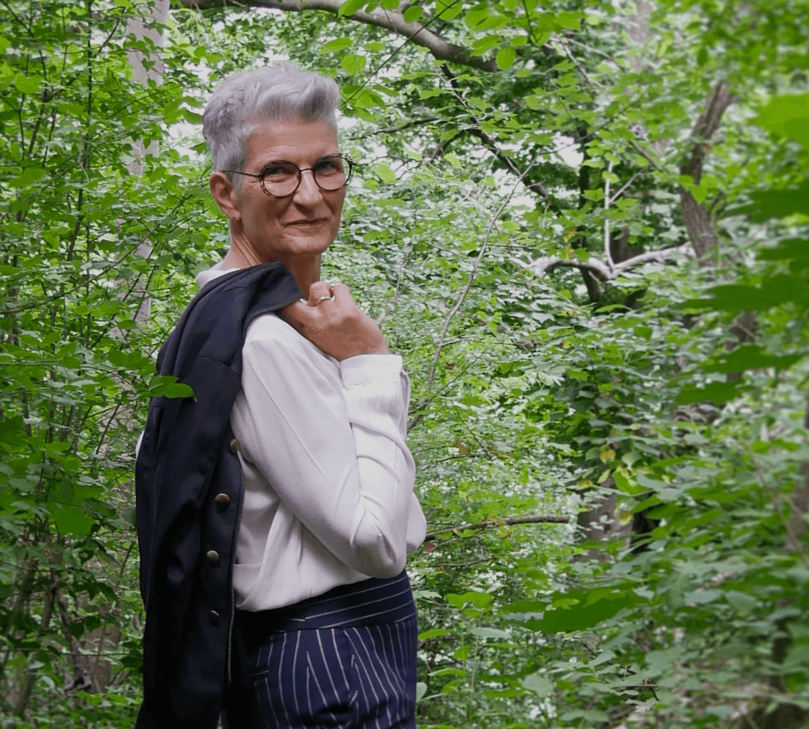 Person mit grauen Haaren und Brille sitzt in einem grünen, bewaldeten Umfeld. Sie trägt ein weißes Hemd und eine dunkelblau gestreifte Hose und lehnt sich mit dem Ellenbogen auf einen Baumstamm, während sie ruhig in die Kamera blickt.