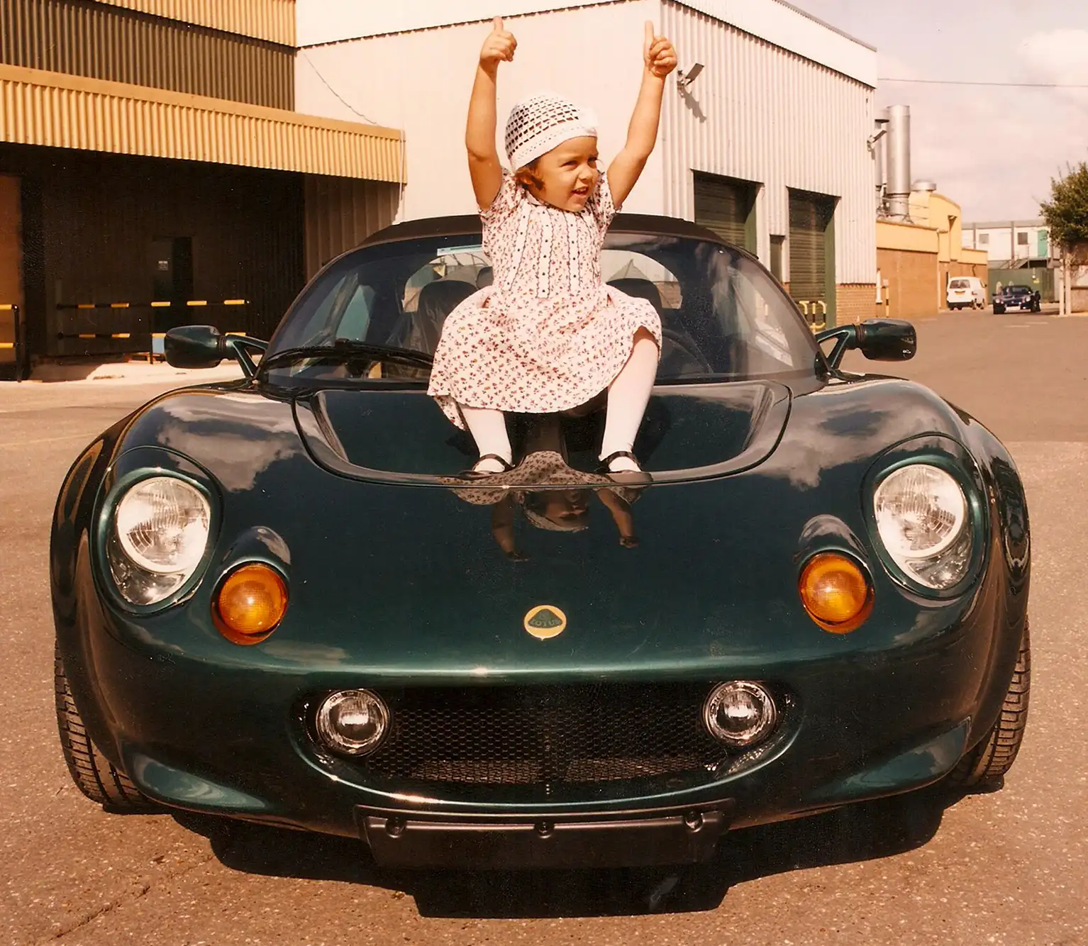 A young Elisa Artioli in a patterned dress and hat sits on the hood of a dark green Lotus Elise sports car, smiling and giving two thumbs up in an industrial area.