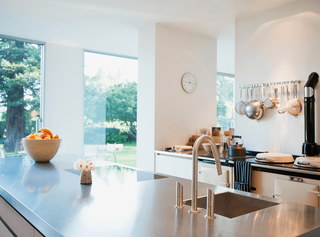 A kitchen with a stainless steel countertop looking out into a bright room with floor to ceiling windows.