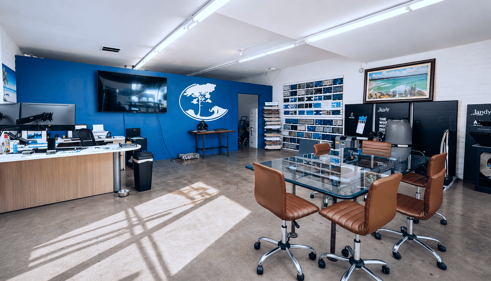 Interior of a small retail or service office space with polished concrete floors, a blue accent wall with a branded globe logo, display shelving, a glass-top conference table with leather rolling chairs, and computer workstations along the perimeter.