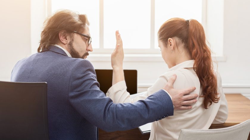 A man in a blue suit and glasses sits beside a woman with red hair in a white blouse, appearing to console her in a bright office setting with a laptop in the background.