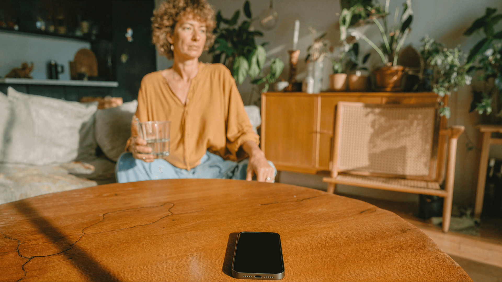 Mulher de cabelos cacheados segurando um copo d'água e olhando para longe, com um celular apagado sobre a mesa à sua frente, ilustrando o abandono de agendamento em clínicas e como a Tivita atua para recuperar esse contato com o paciente.