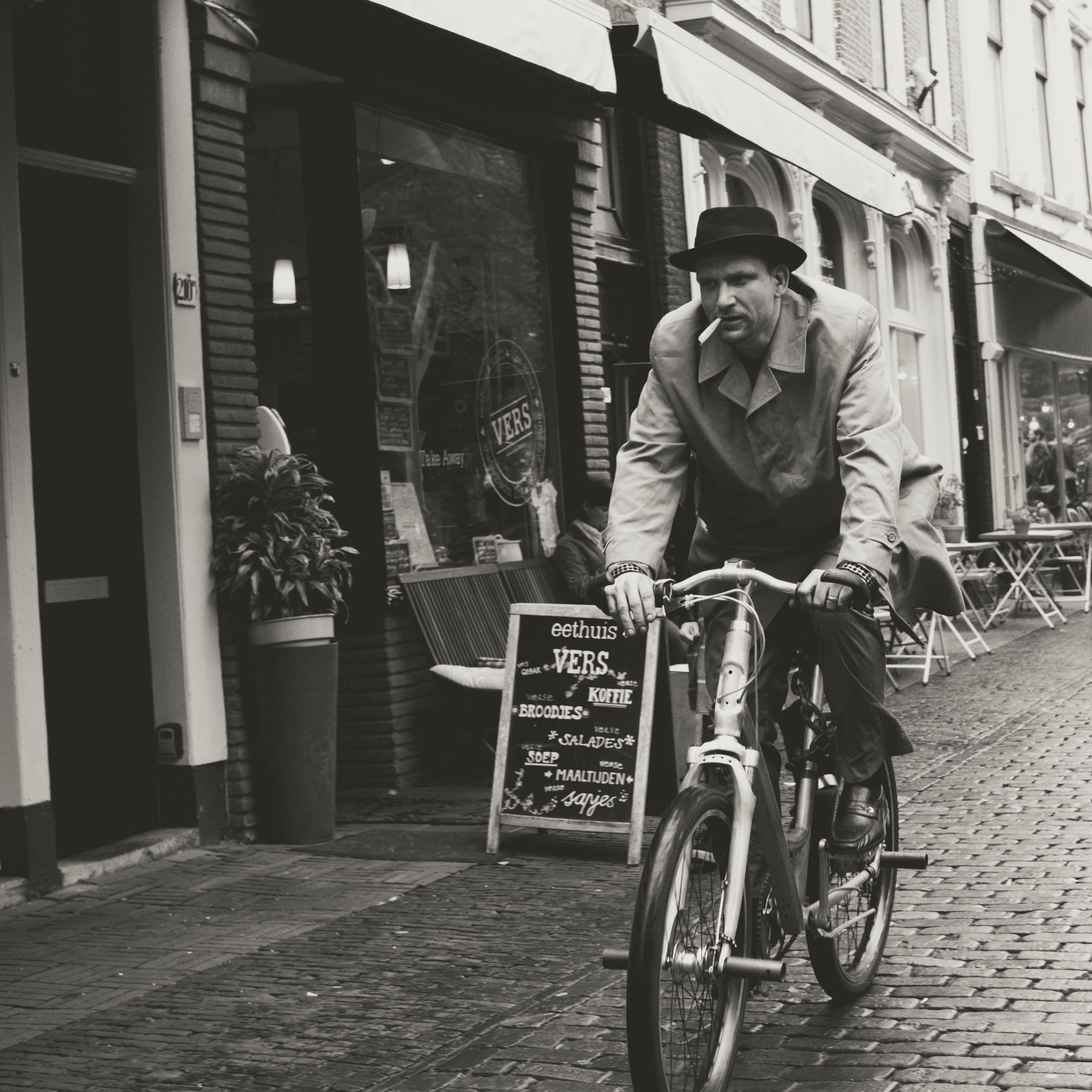 Black-and-white European street photography of a cyclist on cobblestones by a café sign (Eethuis VERS) — ideal for content about Utrecht street scenes, Dutch cafés, urban cycling culture, and vintage-style travel photography.