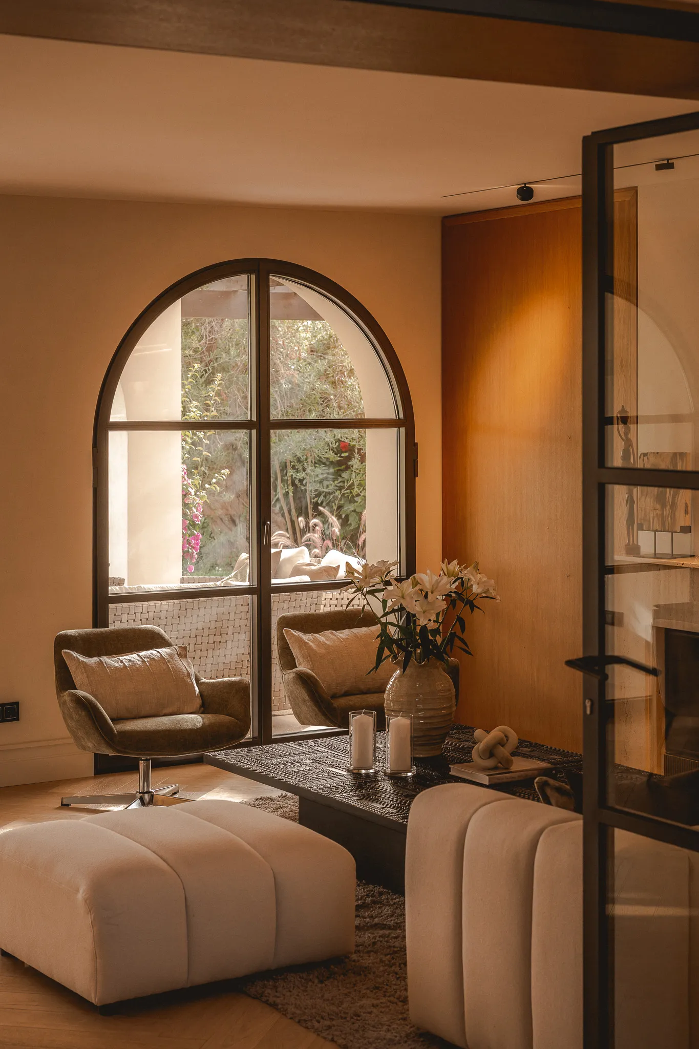 A stylish living room in warm, neutral tones featuring a large arched window in the background, two olive green armchairs, and light-colored modular ottomans in the foreground.