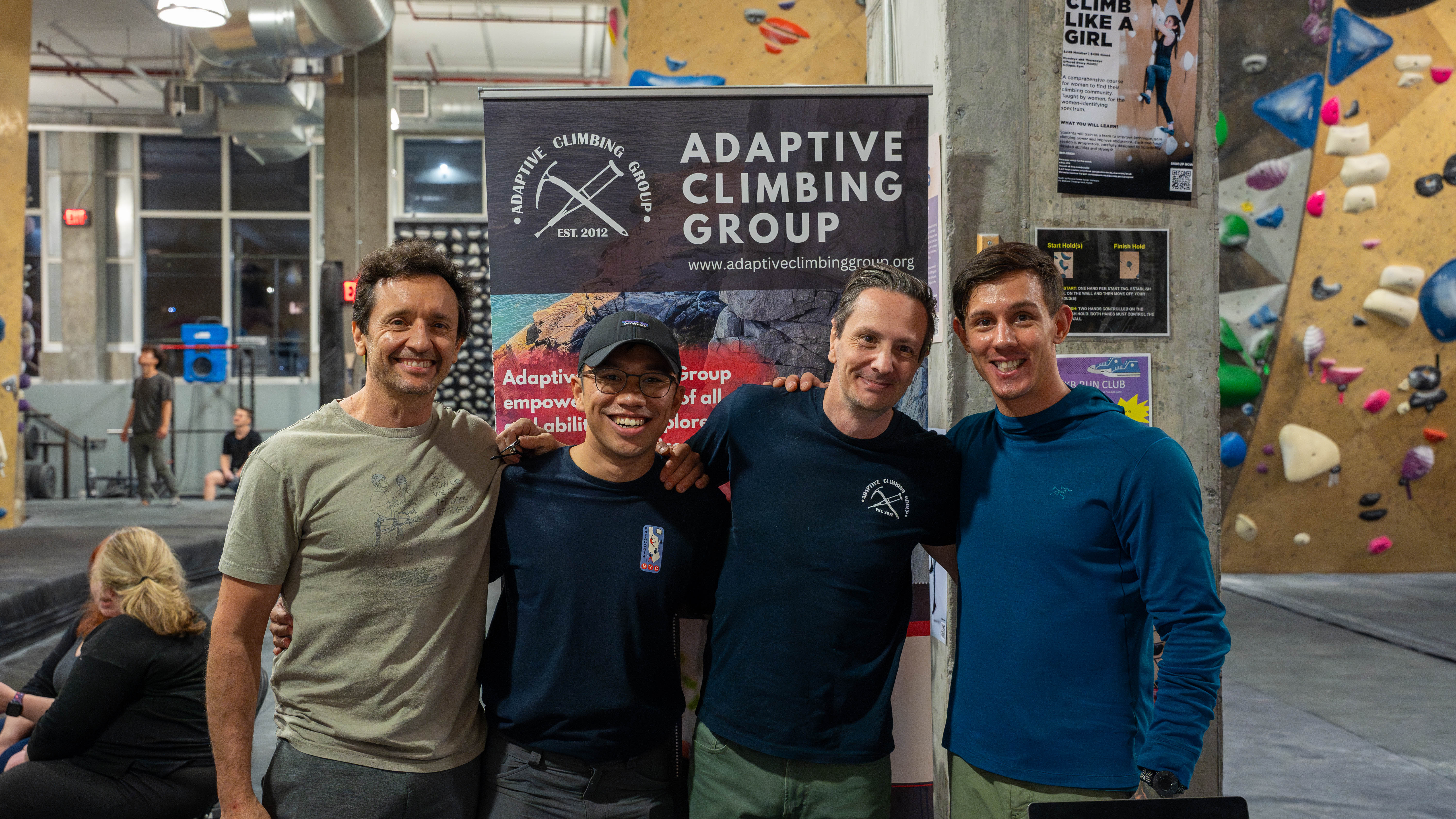 Four gentleman stand smiling at the camera. Behind them a pull up banner reads 'adaptive climbing group'
