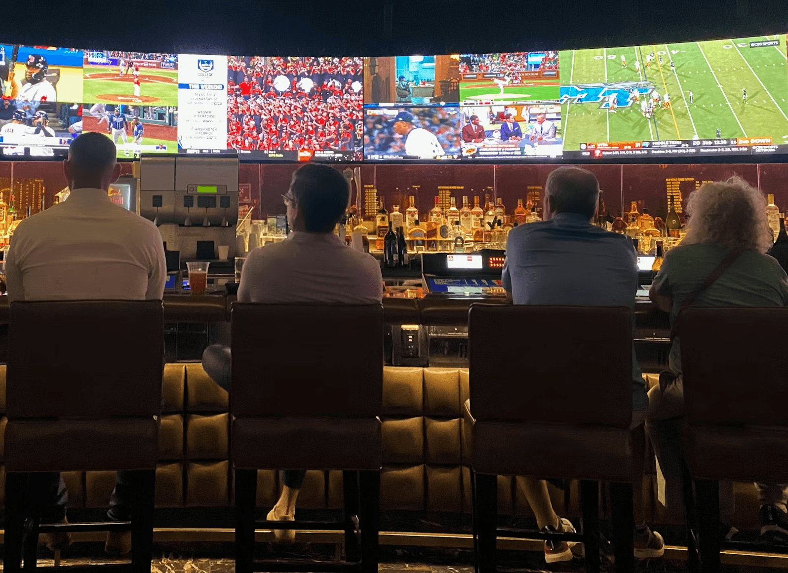 Four people sit at a bar watching sports on a large screen.