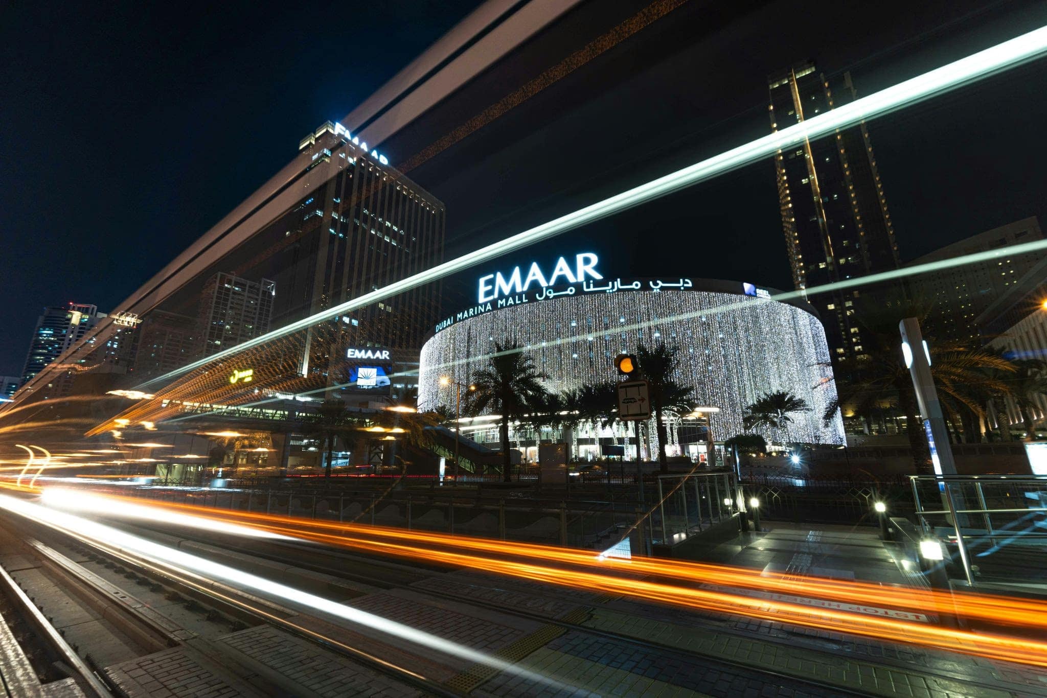 The Dubai Marina Mall at night with illuminated exterior lights, passing tram light trails, and high-rise buildings in the background.