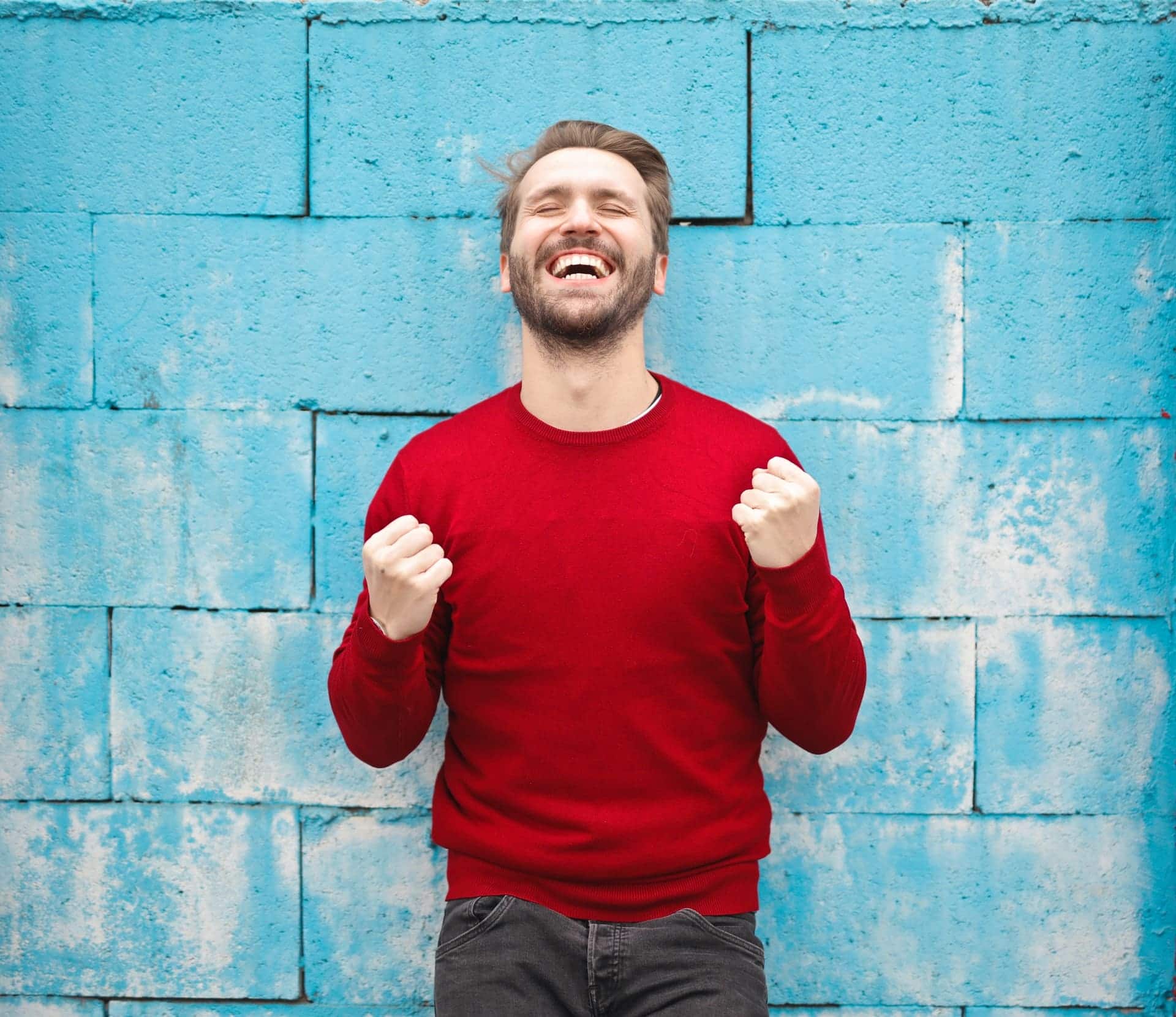 Happy man in red sweater celebrating against blue brick wall.