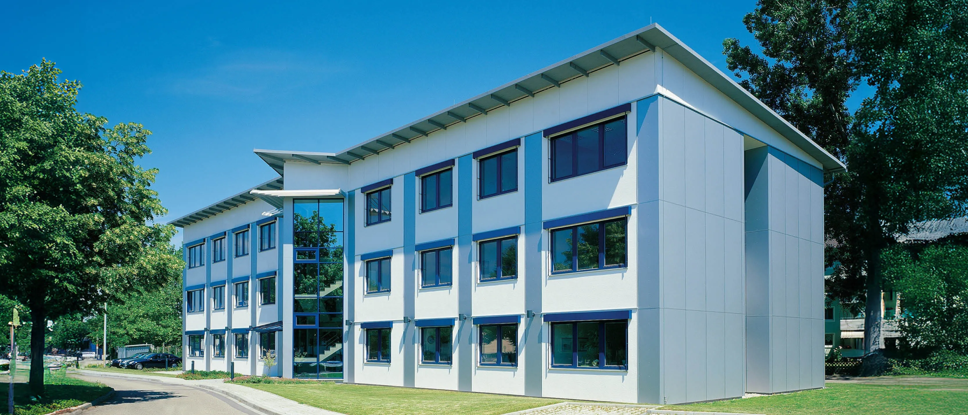 Modernes Bürogebäude mit weißer Fassade und blauen Fensterrahmen, umgeben von Bäumen und Grünflächen.
