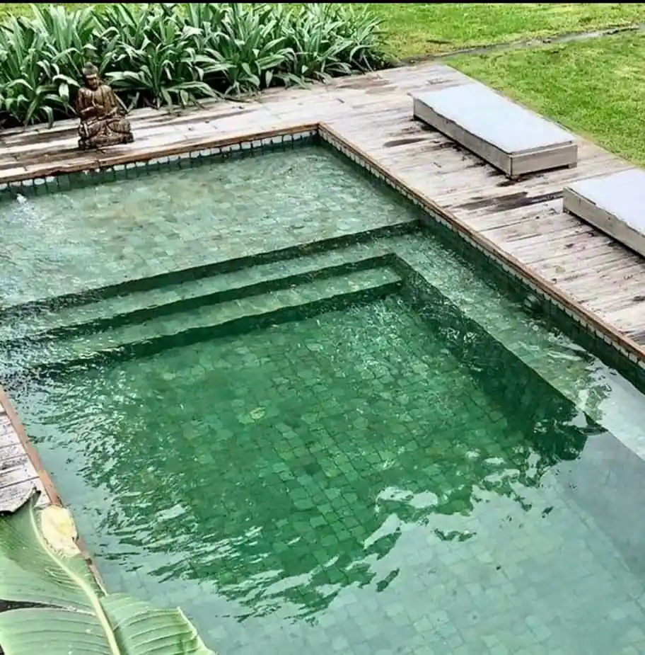 Detalle arquitectónico de piscina premium revestida en tonos verde esmeralda, acompañada de un elegante deck de madera y decoración zen con estatua de Buda.