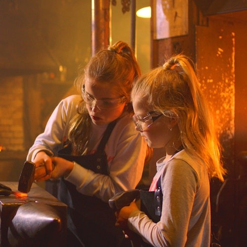 两位年轻女孩戴着安全护目镜和围裙，在灯光昏暗的工作室里敲打金属。