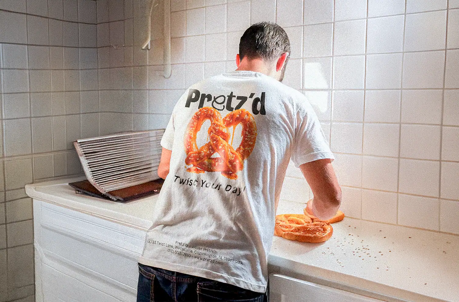 Employé de dos portant un t-shirt blanc Pretz'd avec visuel pretzel photographique et slogan "Twist Your Day!", en cuisine sur carrelage blanc.