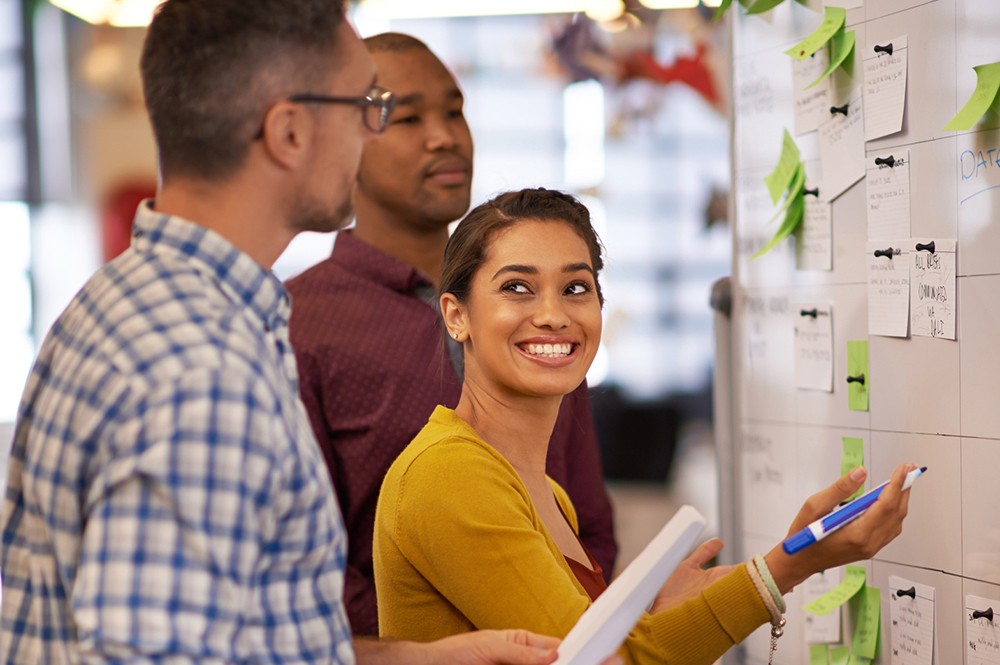 Three colleagues smiling and reviewing notes pinned to a whiteboard during a collaborative planning session.