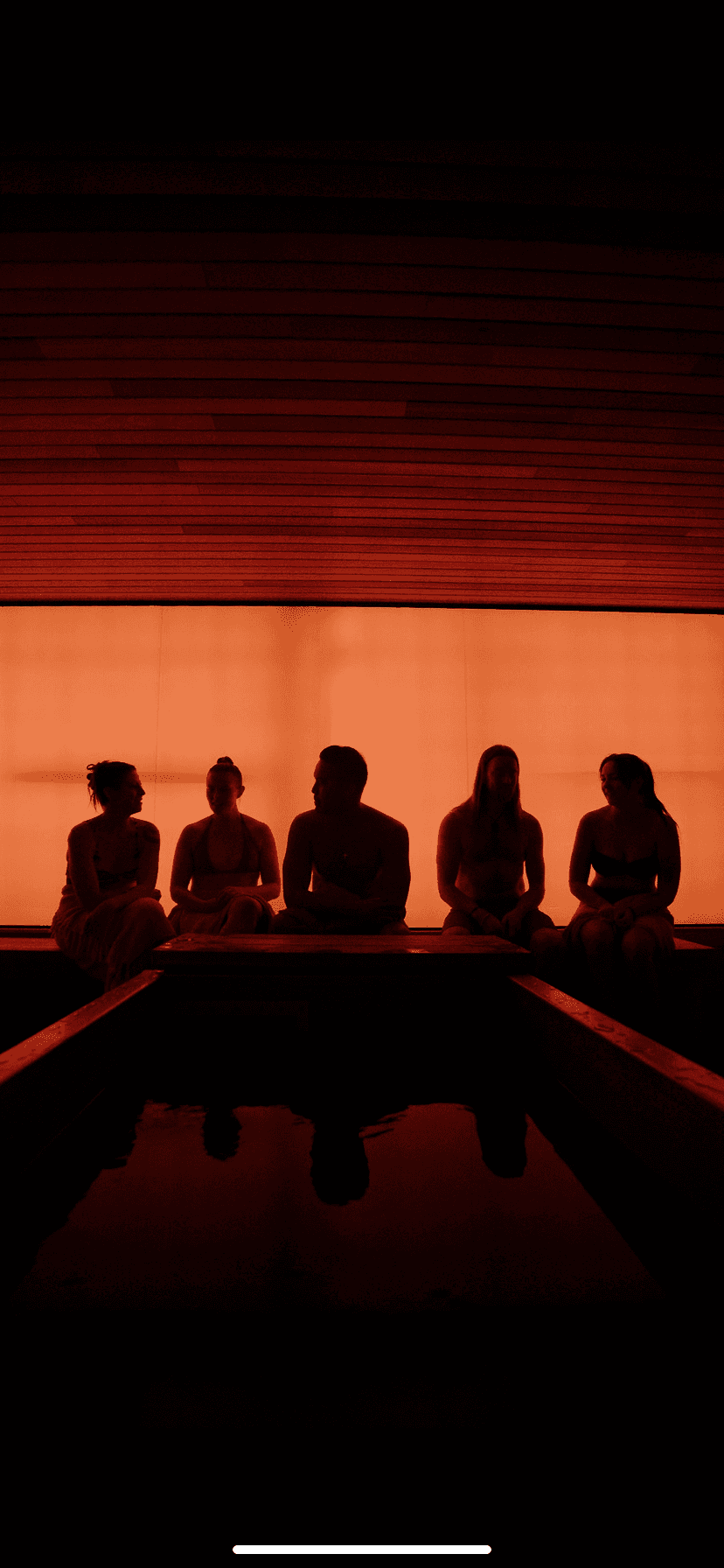 Wooden sauna cabin and hot tub set among rocky mountains and greenery under clear sky.