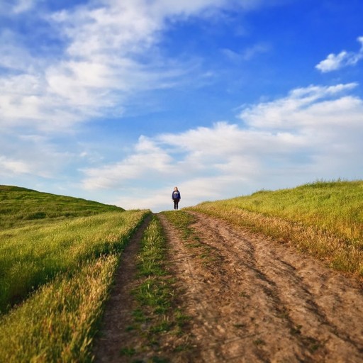 A scenic landscape featuring a dirt path winding through rolling green hills under a bright blue sky with scattered clouds. The path stretches into the distance, lined by grassy fields, creating a sense of openness and tranquility in the countryside.