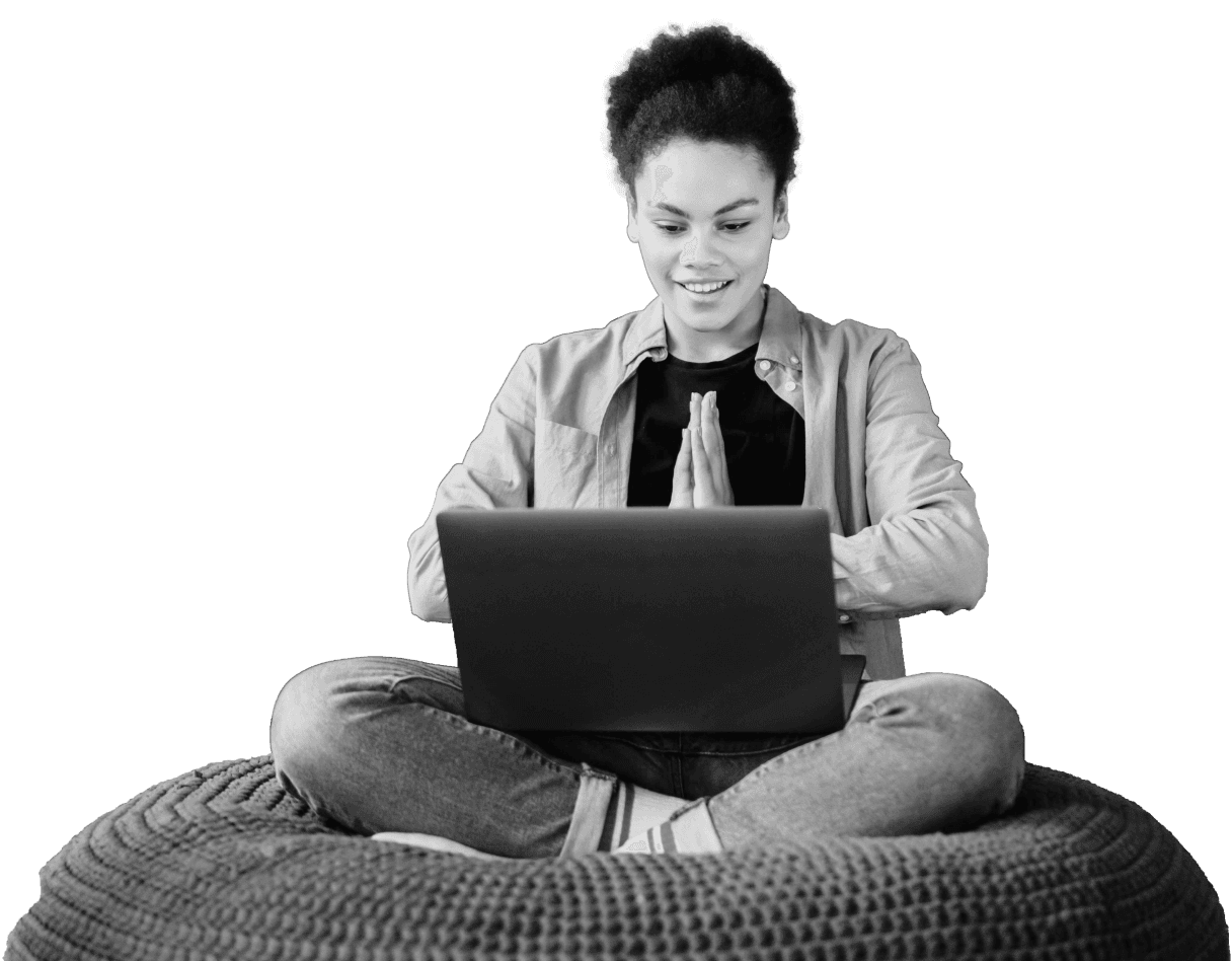 A person sits cross-legged on a pouf, smiling while focused on a laptop screen, suggesting an engaging experience on how to learn trading online.