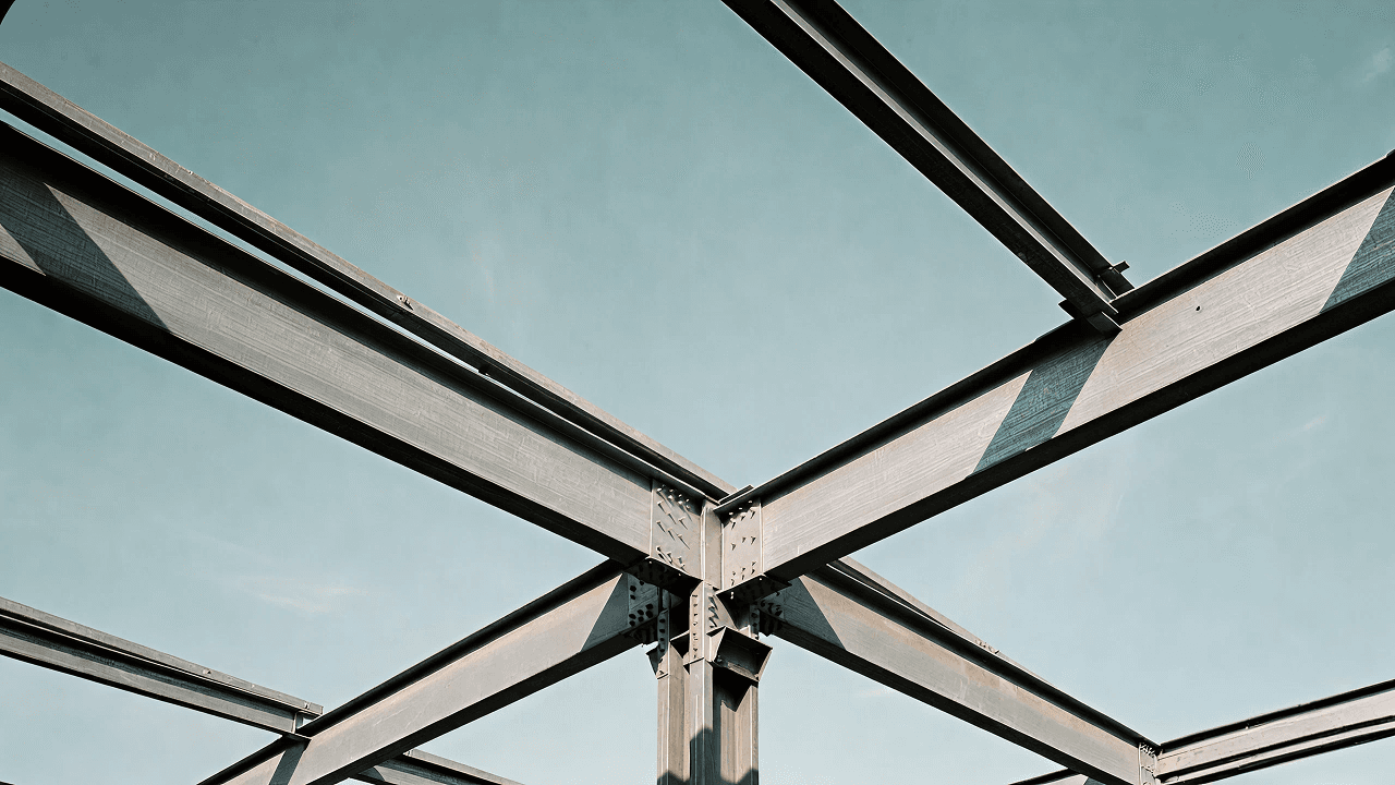 Close-up of crisscrossing steel beams meeting at a bolted structural joint, viewed from below against a clear sky.