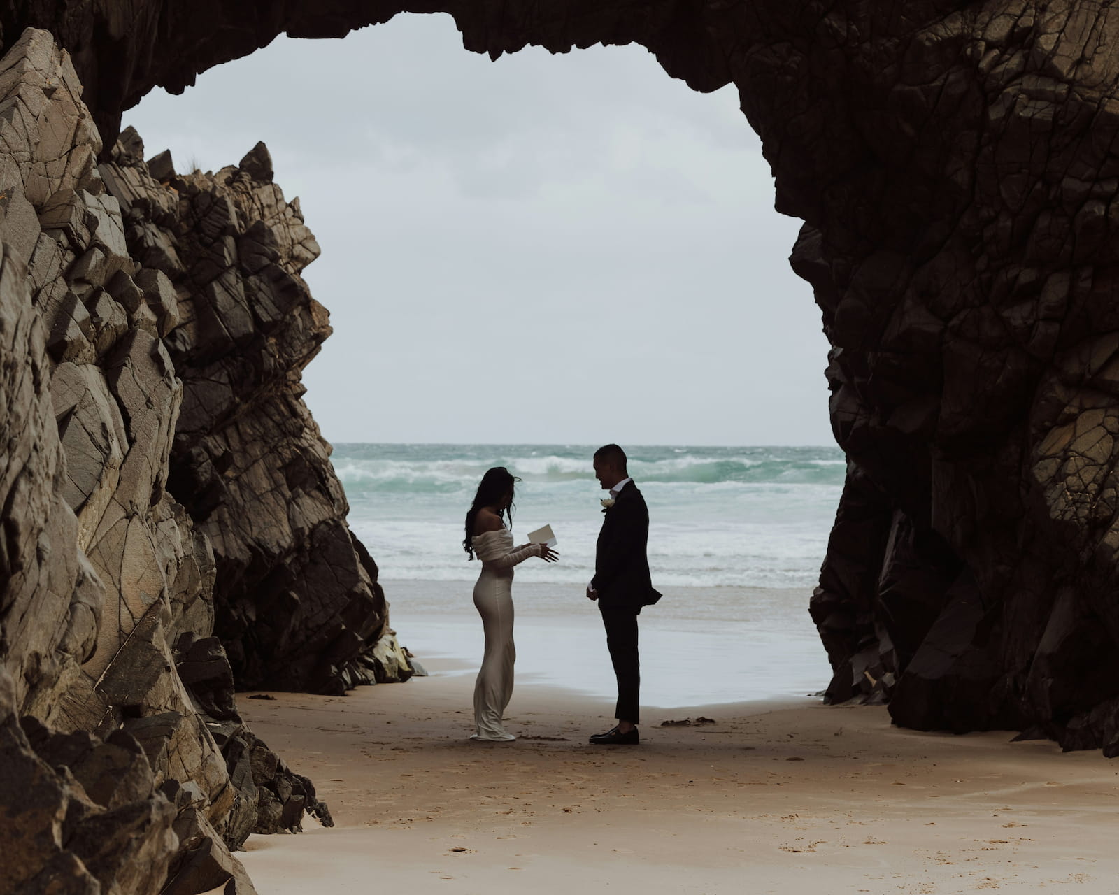 Couple reading their vows to each other framed by a natural rock arch on a sandy beach with ocean waves in the background