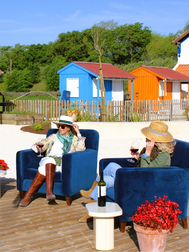 Two people relax in blue chairs outdoors, sipping wine. Vibrant blue and orange beach huts are in the background, with a sunny, cheerful atmosphere.