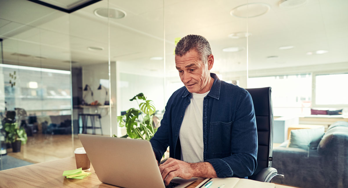 Man working on a laptop in a modern open-plan office with sticky notes and a coffee cup nearby