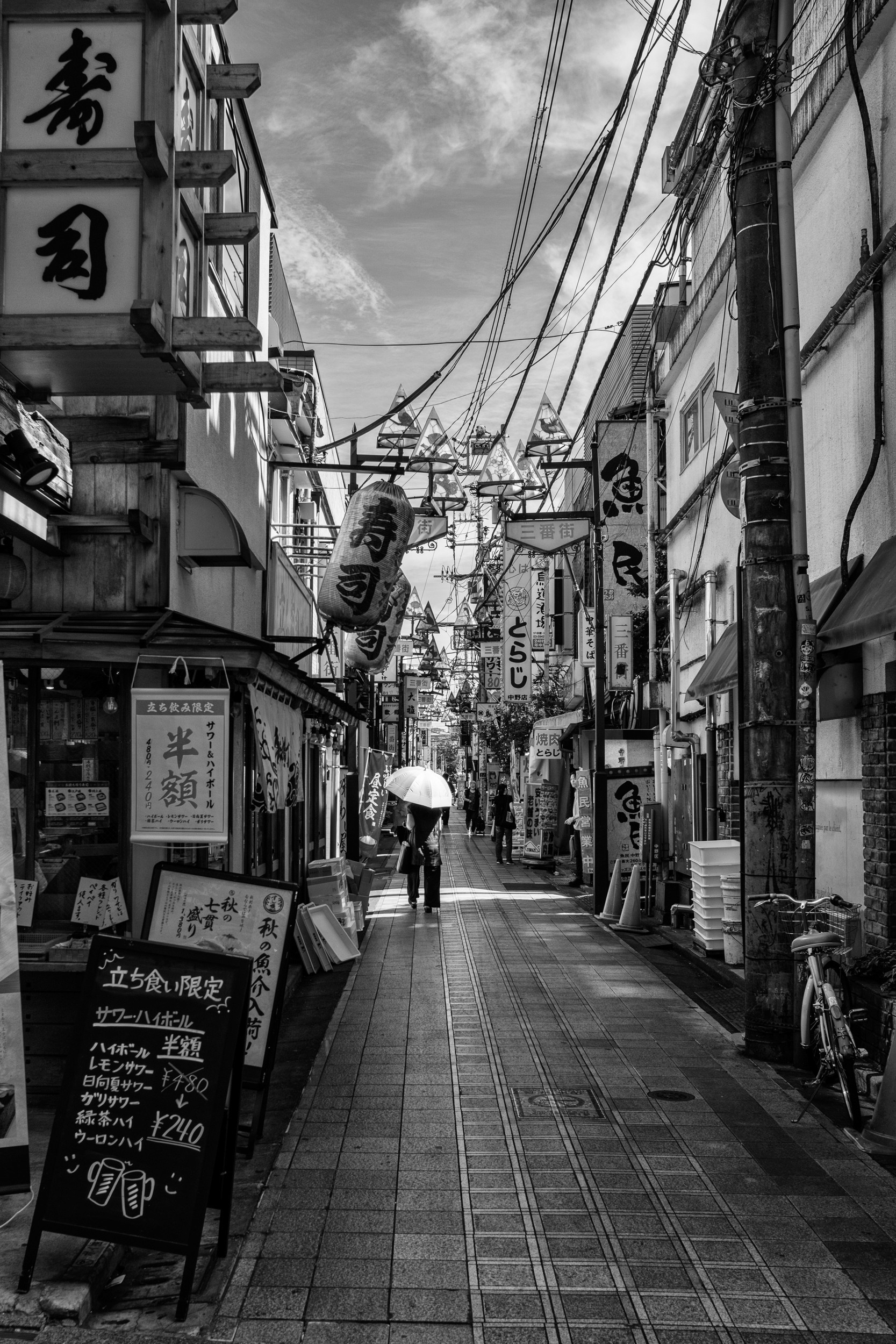 Black and white street photograph of a person holding an umbrella walking down a narrow shopping street.