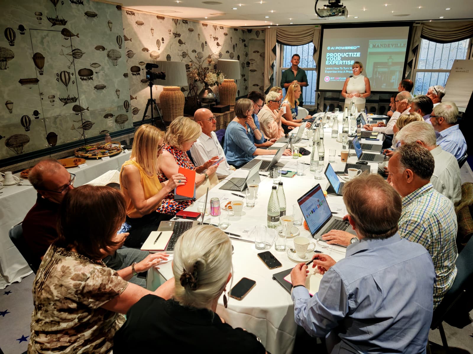 Cozy reading nook with sofa, chair, and plants inside a glass cabin with mountain forest views.