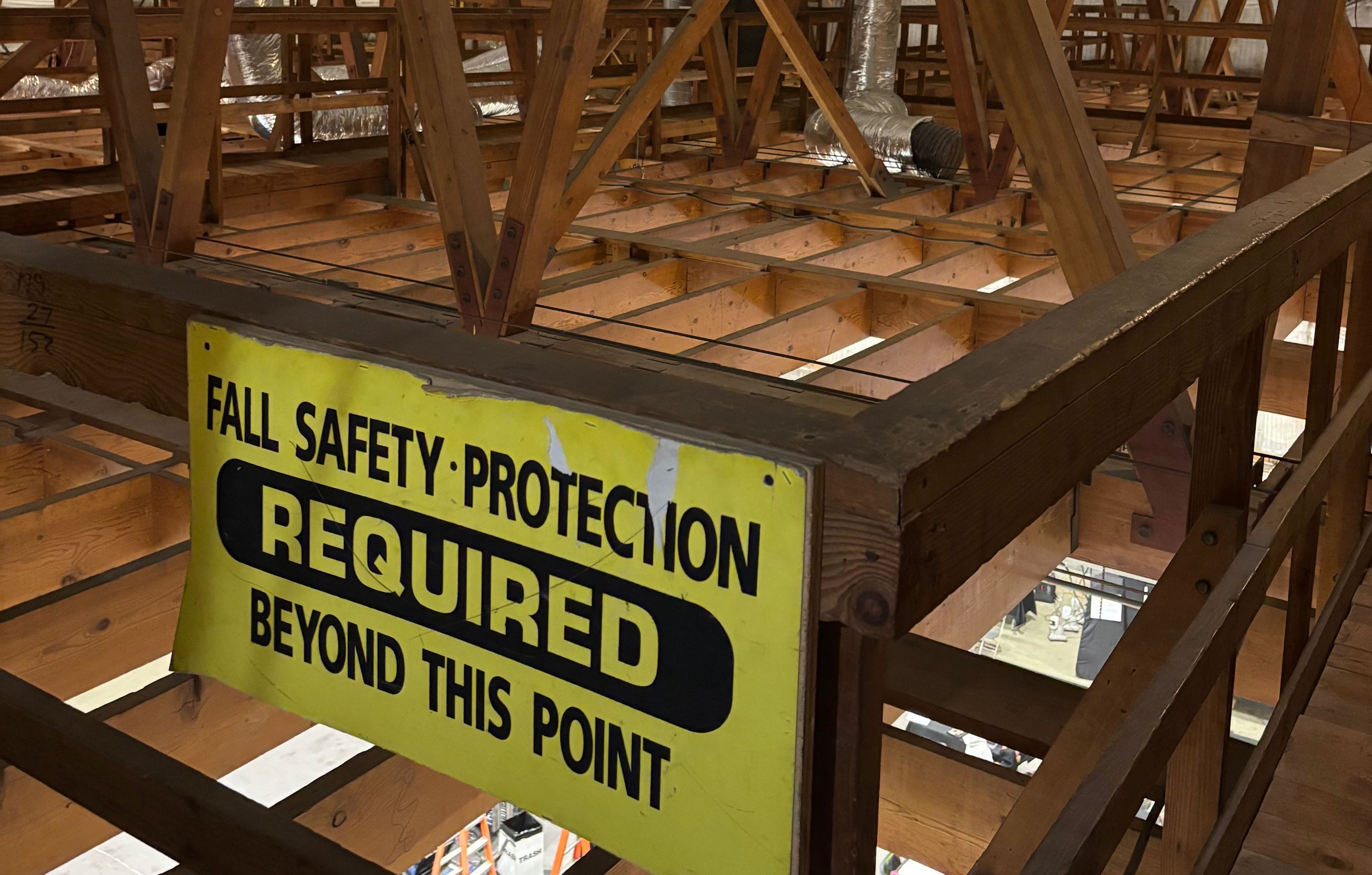 A yellow ‘Fall Protection Required Beyond This Point’ sign mounted on the wooden perms above a stage.