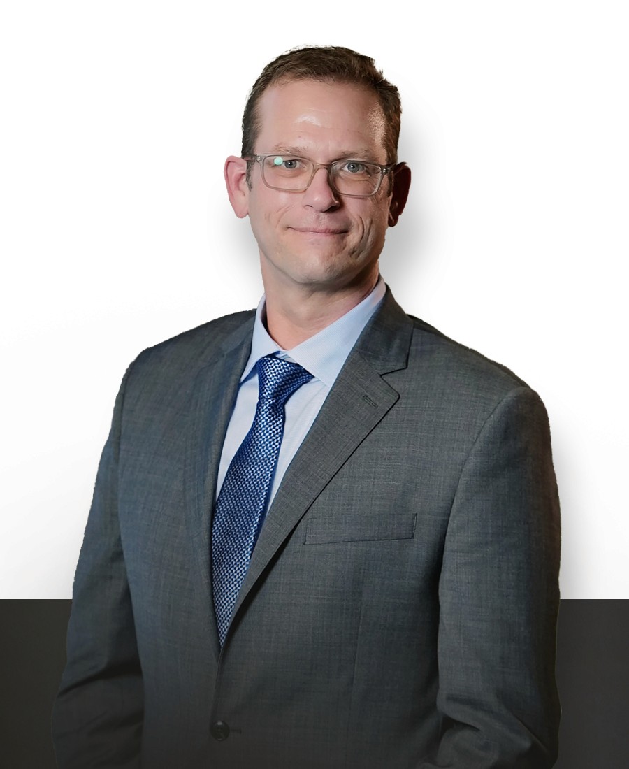 corporate headshot male with light skin and light brown hair wearing glasses in a grey suit with light blue shirt and dark blue patterned tie with a closed-lip slight smile. 