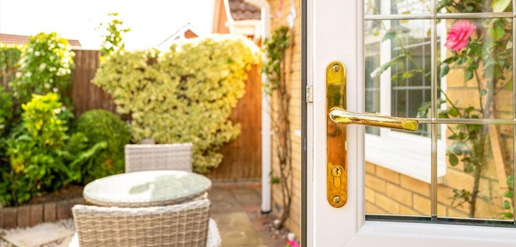 An open white patio door with a golden lock, leading to a sunlit patio with lush surroundings.