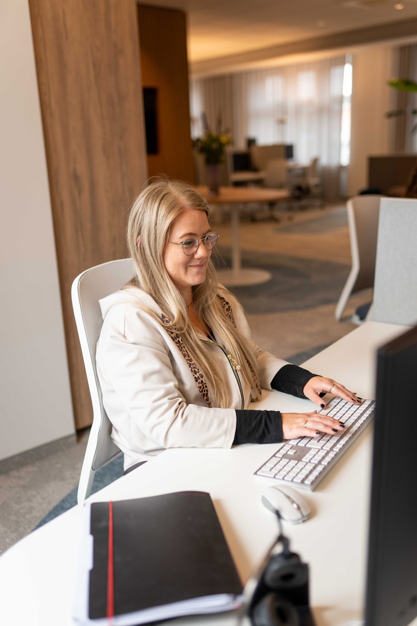 Mitarbeiterin im hellen Blazer sitzt vor ihrem PC an einem Tisch im Büro von SLB.