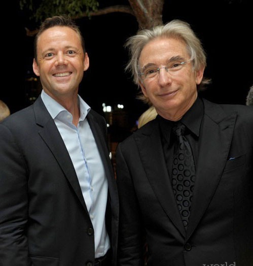 A man in a blue suit and a conductor all in black smile after a performance.