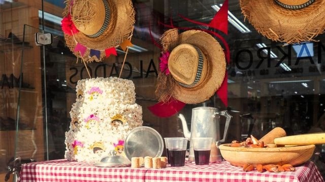Vitrine de São João com comidas típicas sobre mesa com toalha xadrez vermelha; chapéus de palha pendurados no vidro ao fundo.